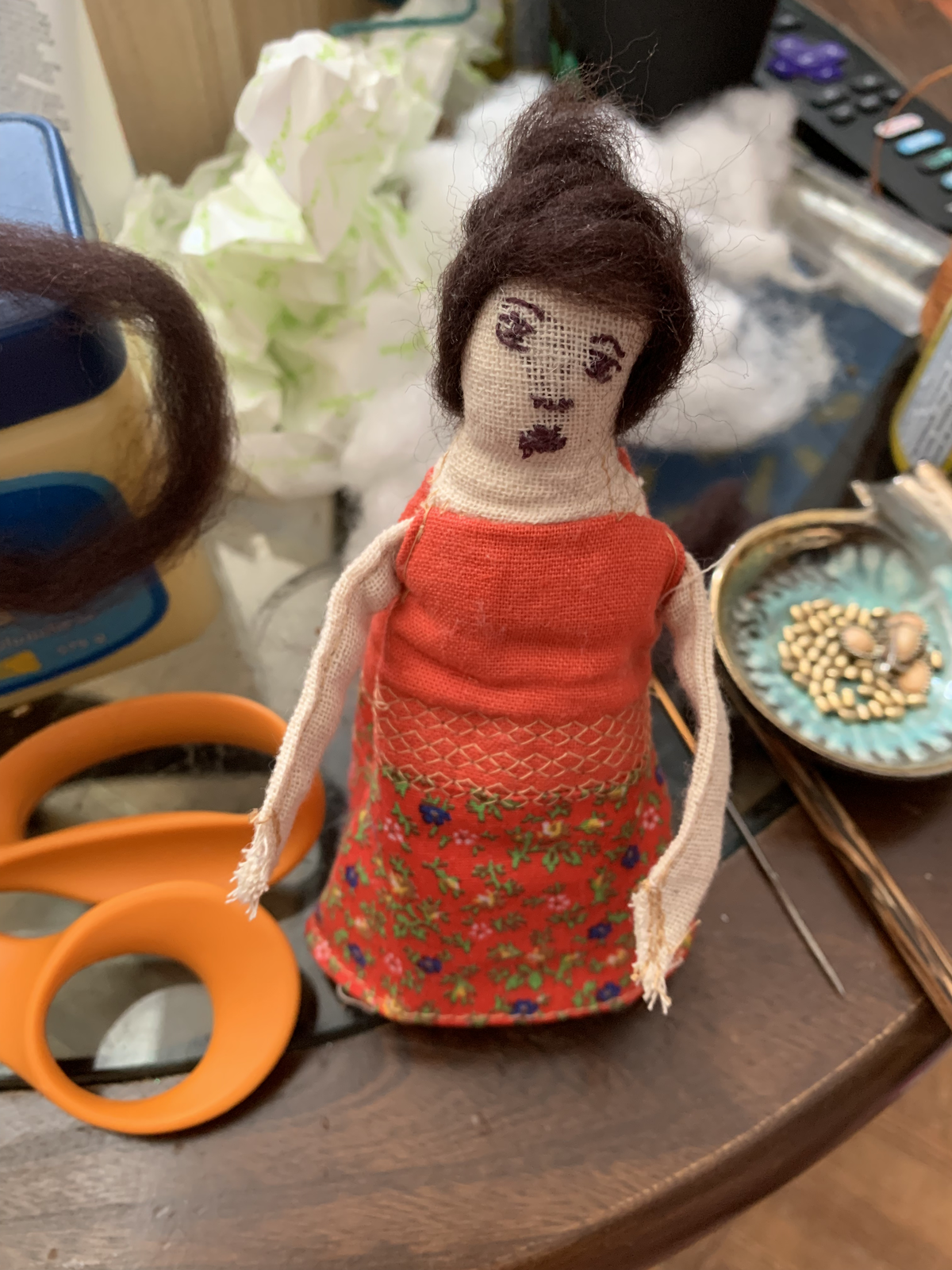 Genealogy doll in red floral dress on worktable — embroidered face, dark wool hair in updo