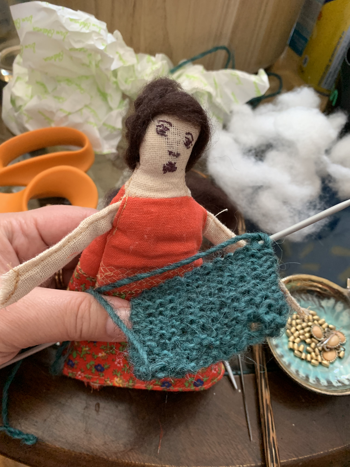 Genealogy doll in red dress with teal knitted shawl being made — knitting needles and yarn