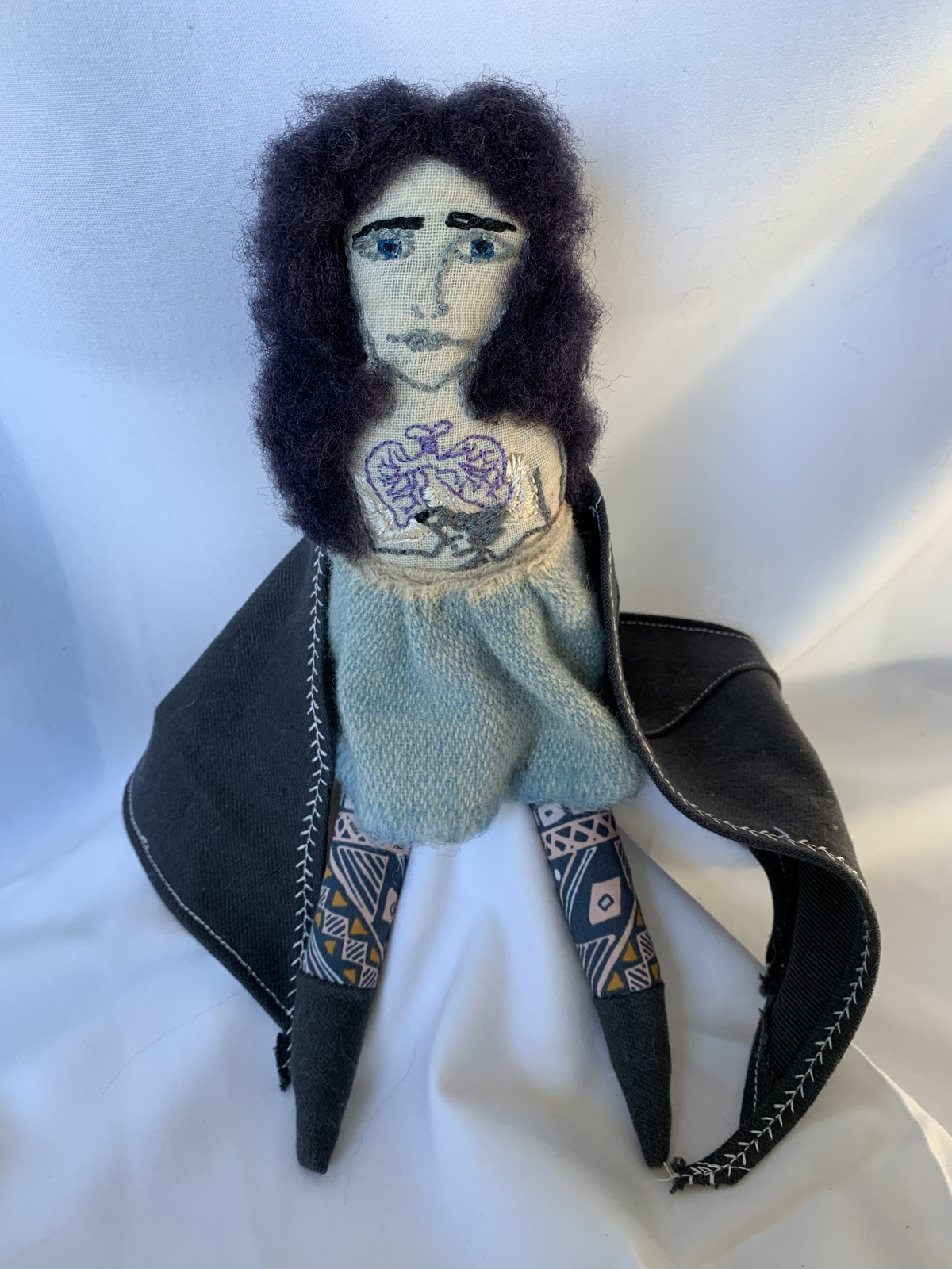 Genealogy doll with dark curly hair, embroidered chest tattoo, light blue sweater, dark jacket with white stitching, geometric print leggings