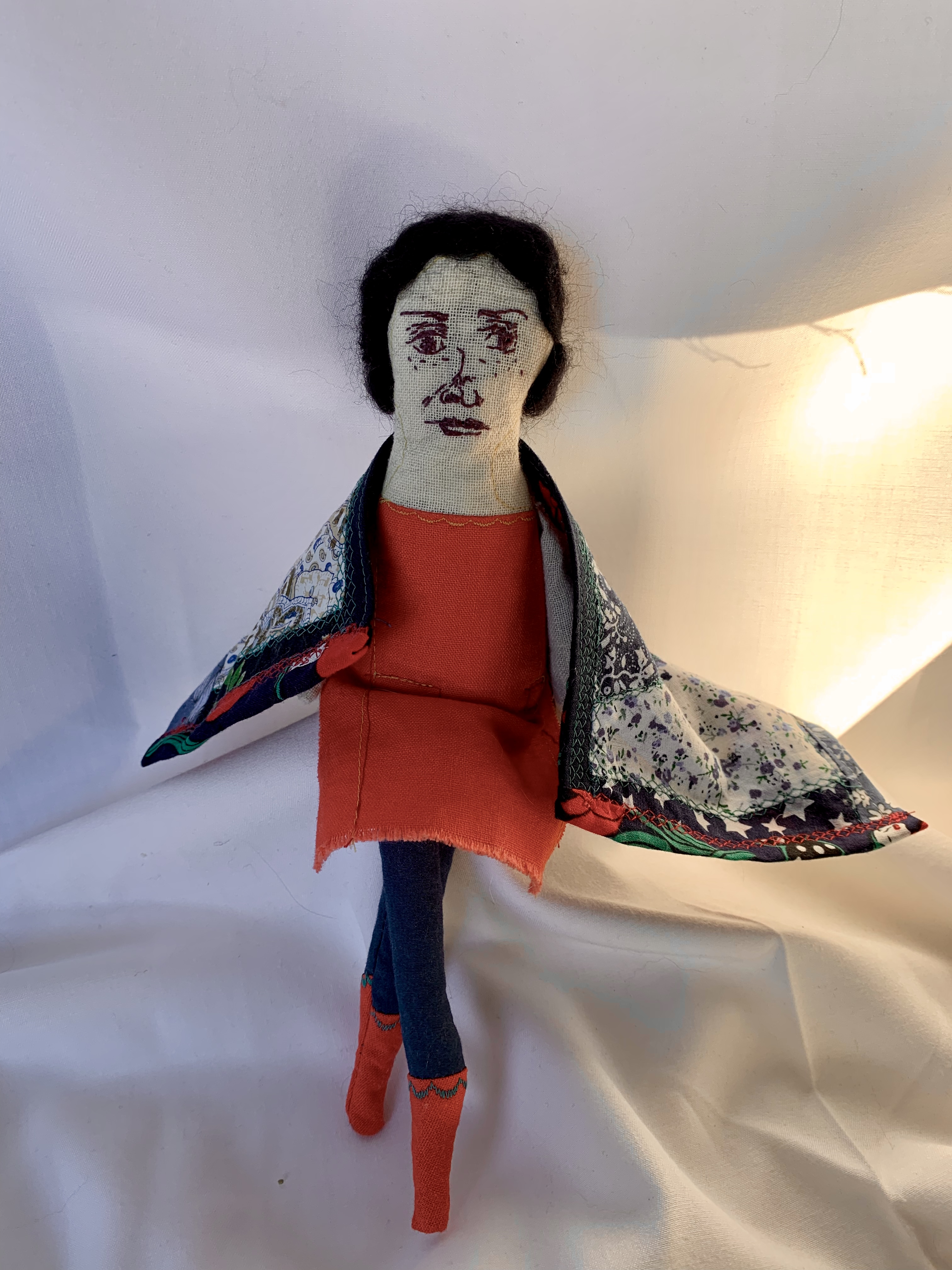 Genealogy doll in terracotta dress with patchwork floral shawl, arms spread wide, navy leggings and coral socks