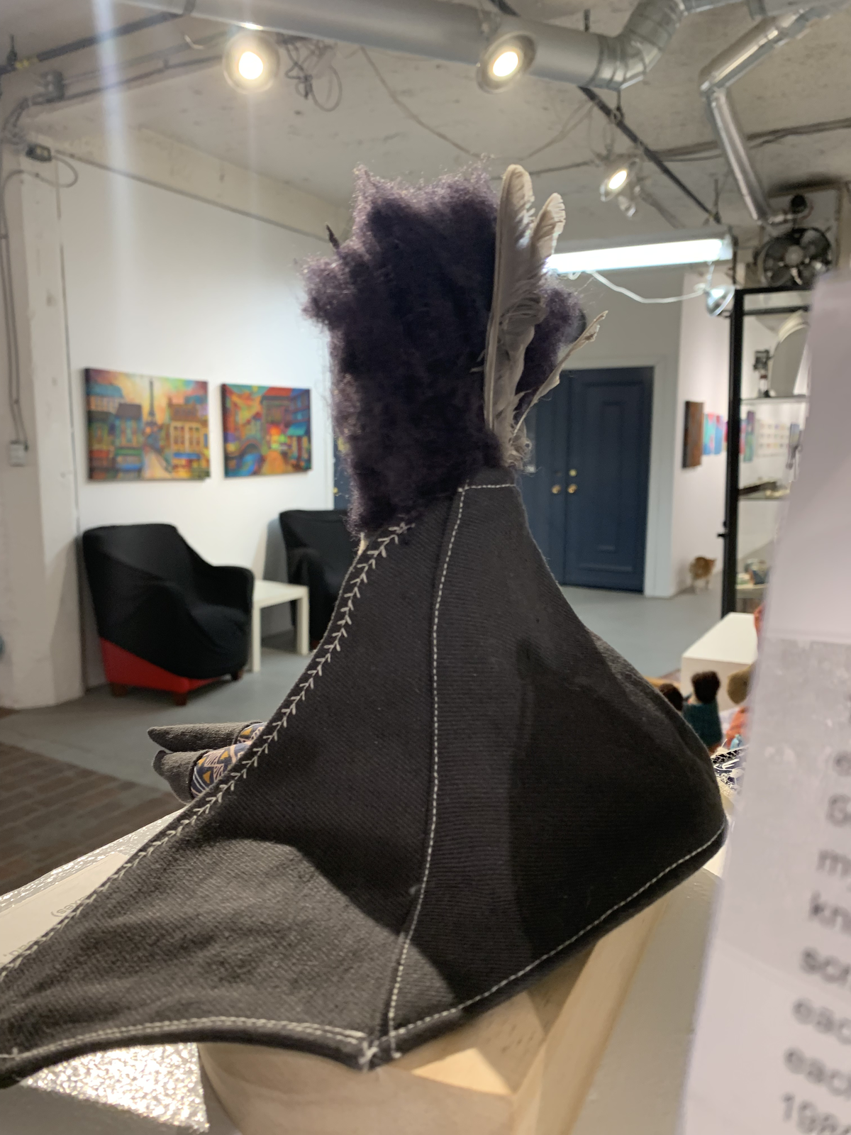 Euphrosine — back view showing dark jacket with white blanket stitching and feather in hair, gallery setting