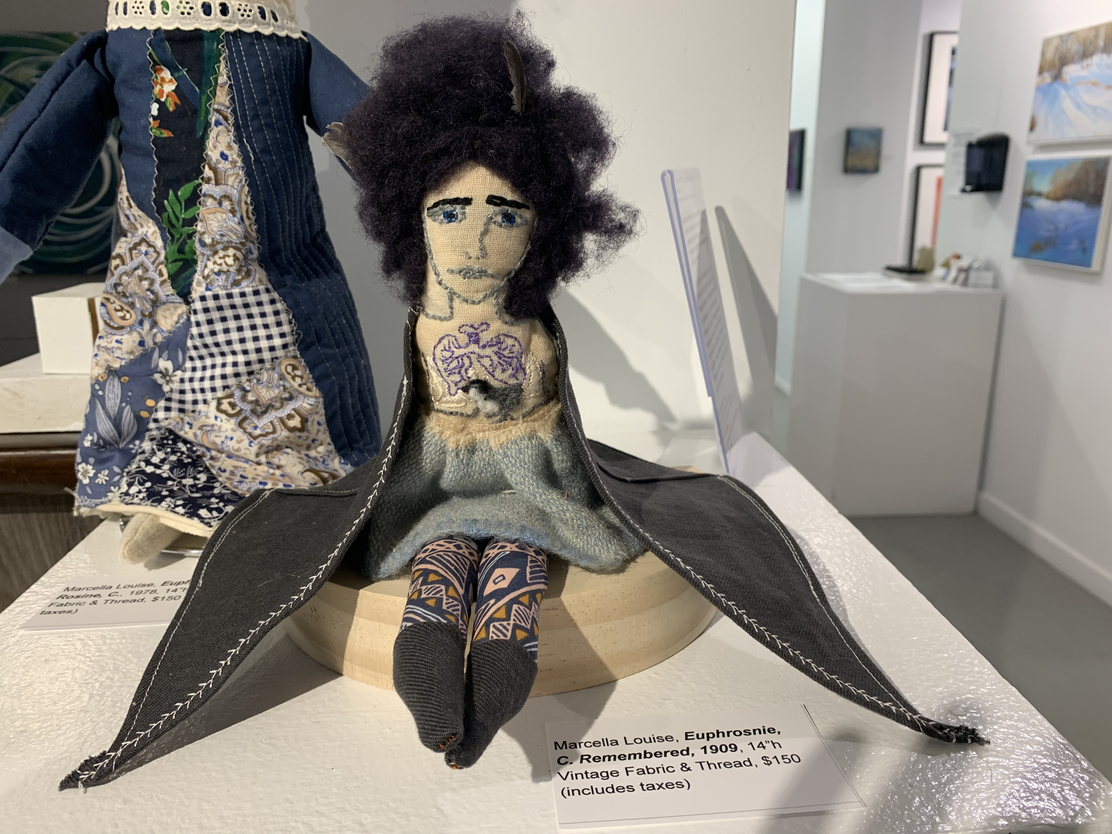Euphrosine — genealogy doll seated in gallery, dark wool hair with feather, embroidered tattoo, dark jacket, geometric print leggings
