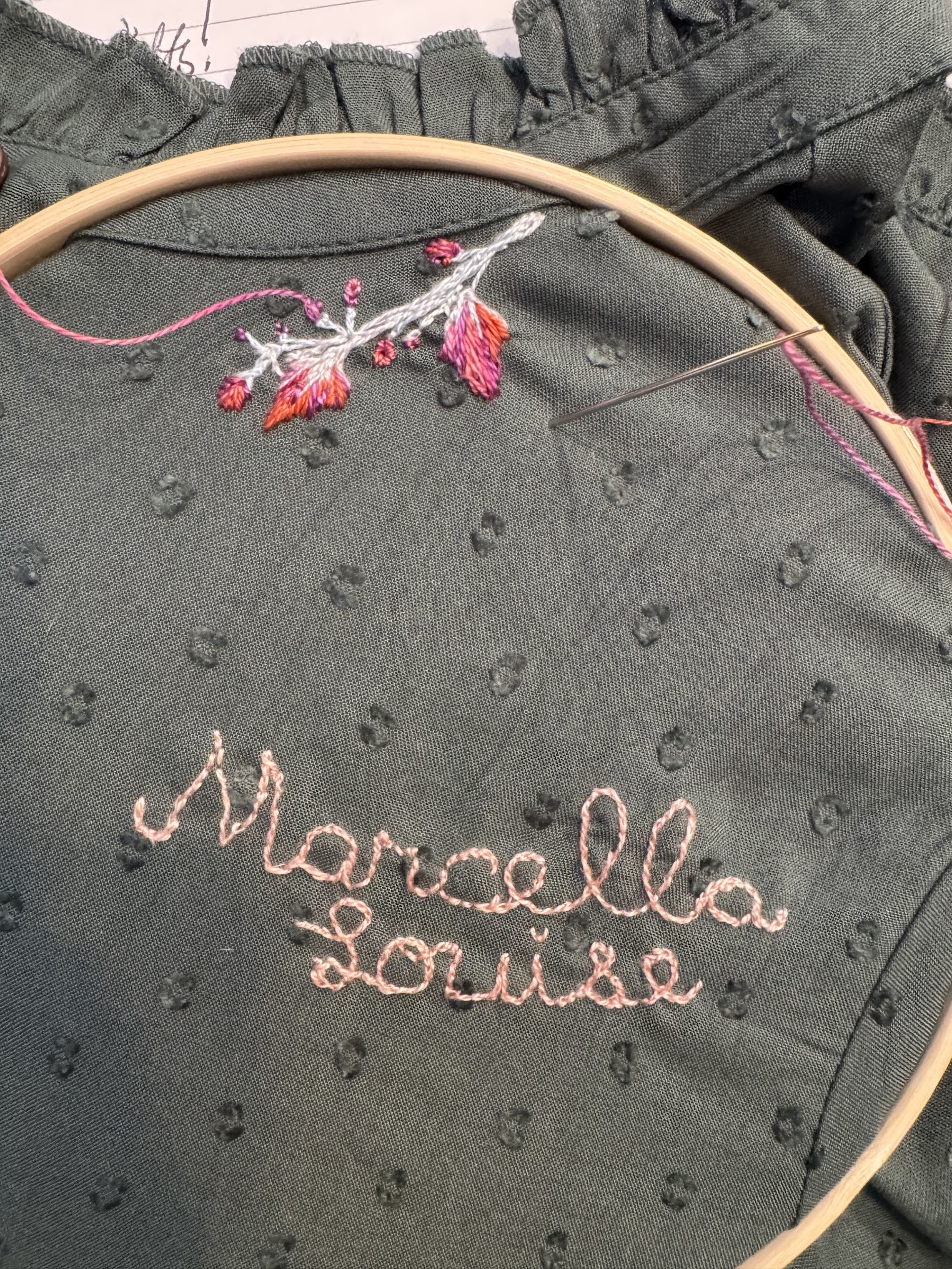 Embroidery hoop on green dotted fabric stitched with 'Marcella Louise' in pink and a floral motif