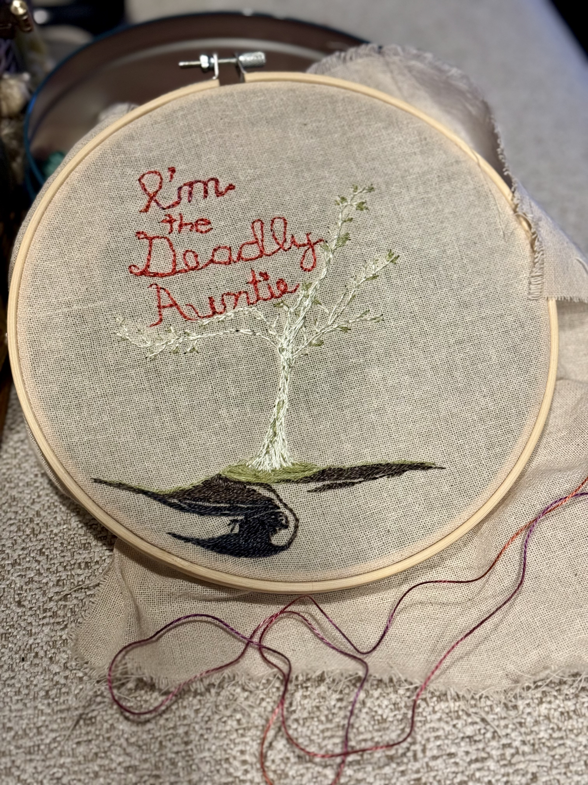 Embroidery hoop on linen — 'I'm the Deadly Auntie' stitched in red with a white tree, green leaves, and a dark bird