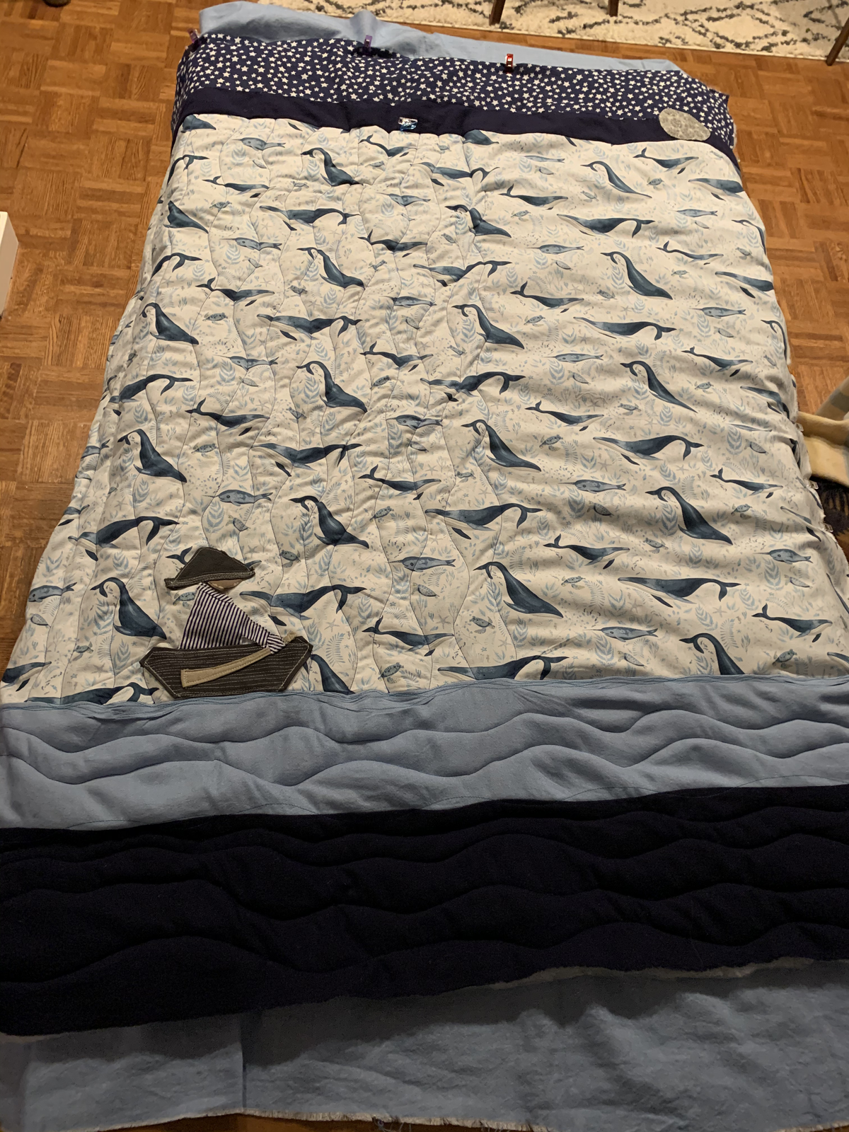 Under the Light of the Moon quilt laid flat — whale print, starry sky, layered blue and navy wave strips at the bottom