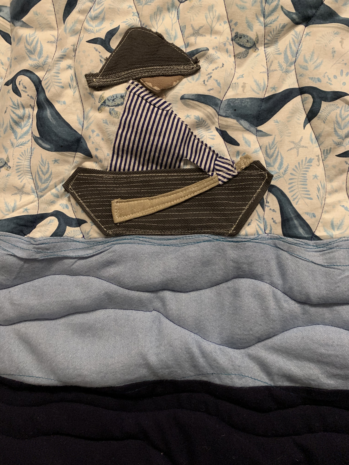 Close detail — appliqué boat with striped figure rowing on layered blue waves, whales and sea turtles swimming behind