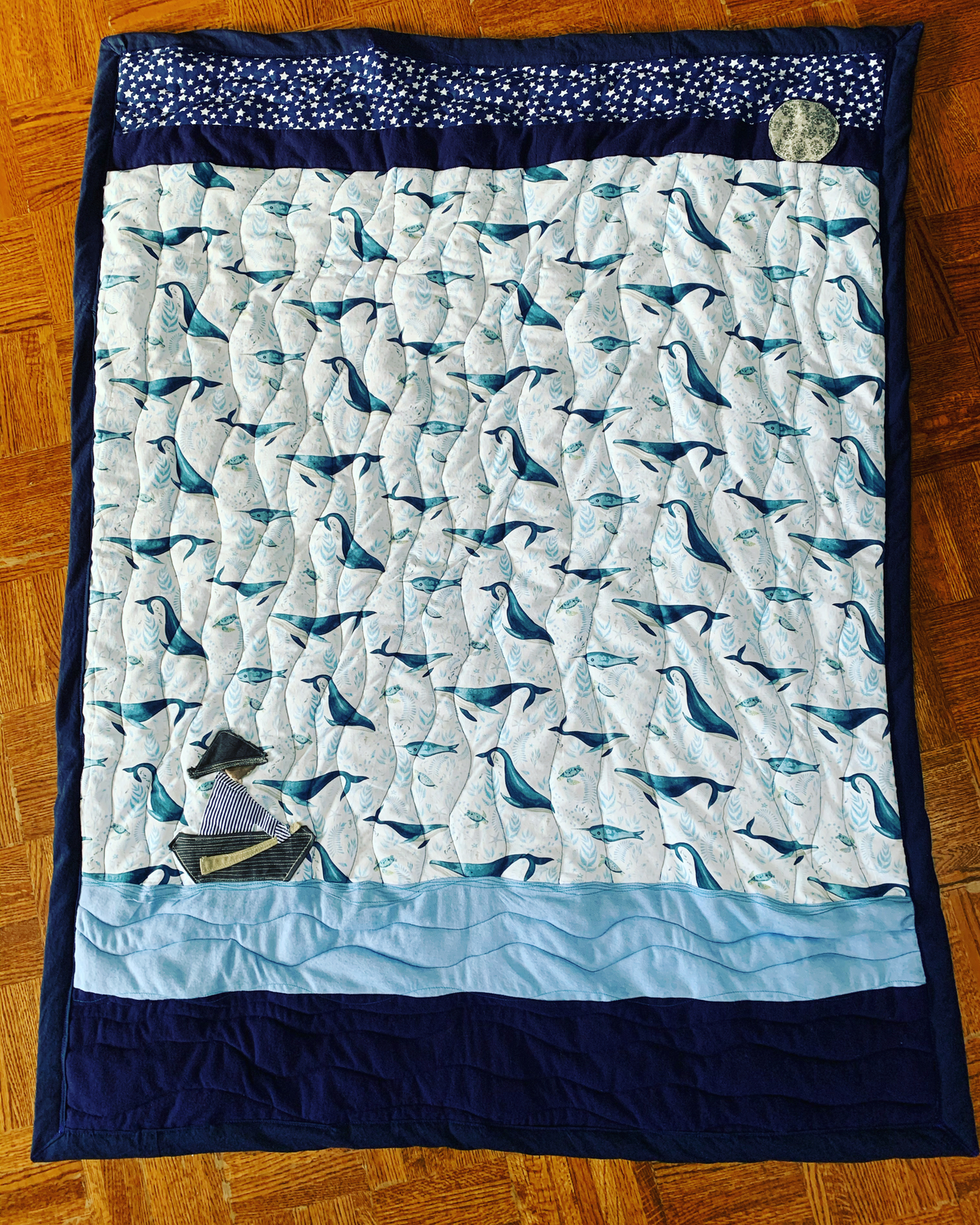 Under the Light of the Moon quilt — whale print centre panel with navy border, starry sky strip, appliqué moon, layered ocean waves, and a small boat with figure