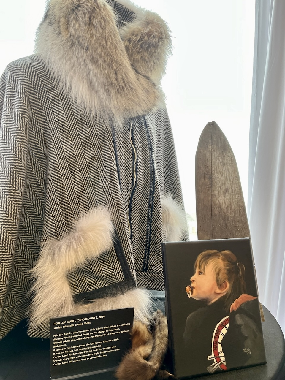 Pchi Loo Auntie exhibition: fur-trimmed herringbone cape on display with artist placard, 2024