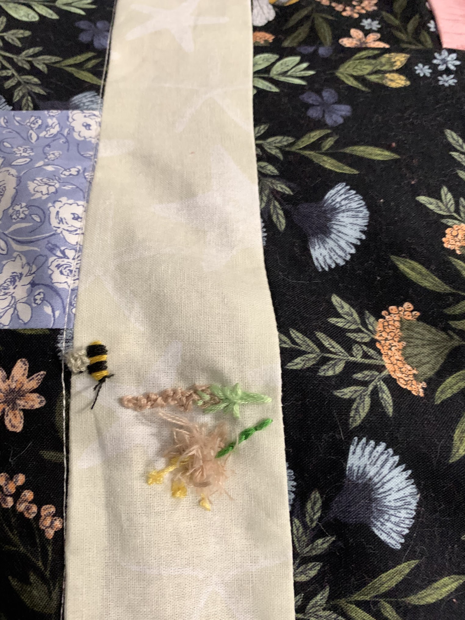Close detail of Practical Magic quilt — a small embroidered bee beside a pale wildflower sprig stitched into the cream sashing