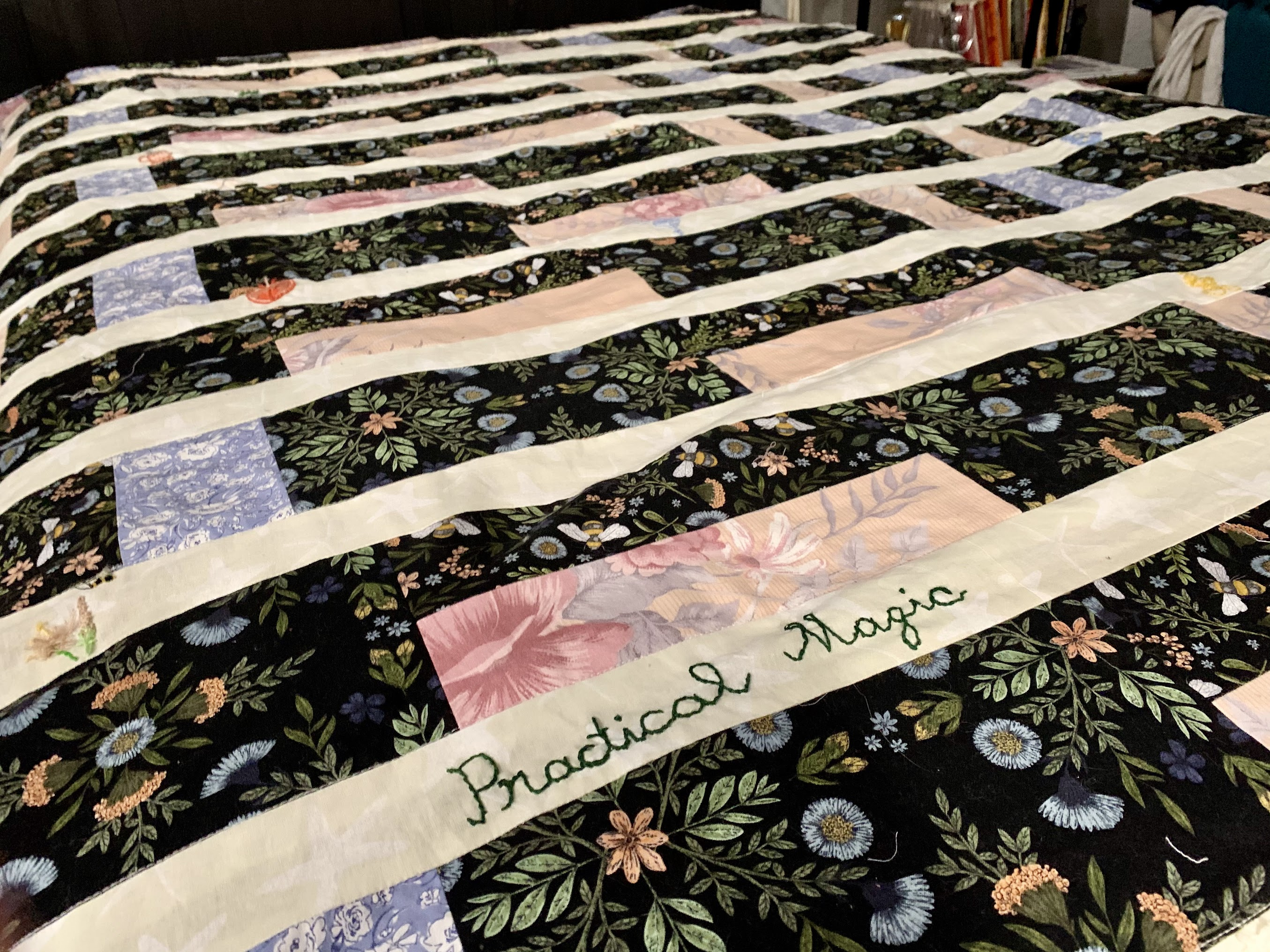 Practical Magic quilt laid out — black botanical print with cream sashing, pink and blue floral inserts, name embroidered in green thread