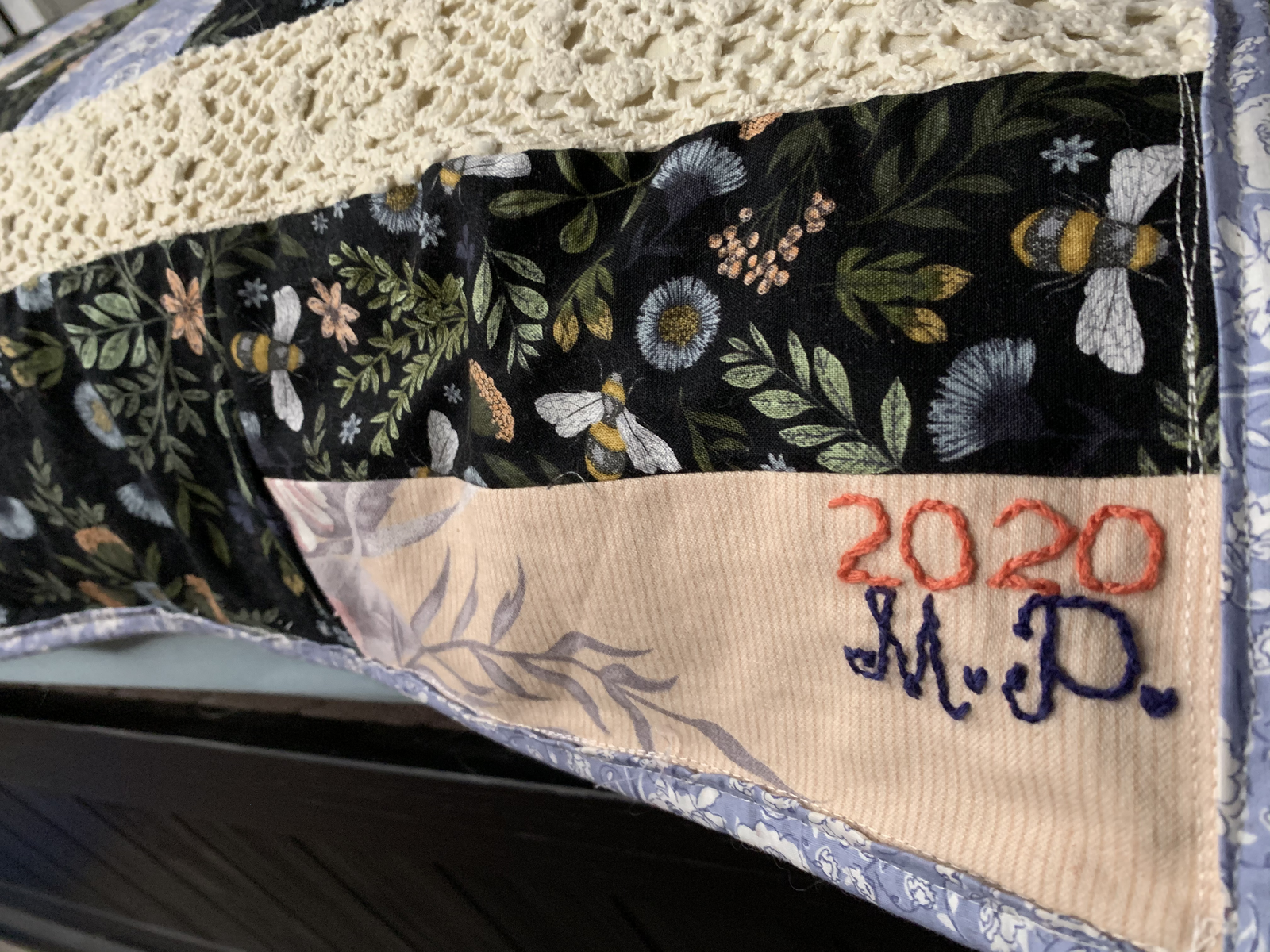 Close detail of quilt label — '2020 M.P.' embroidered in coral and navy thread on cream fabric beside dark floral bee print and crochet lace