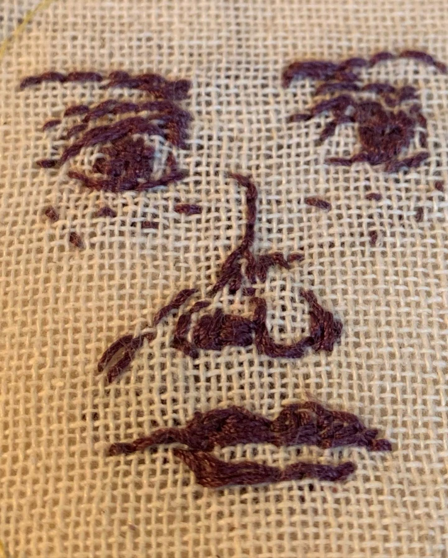 Close detail of a face embroidered in dark aubergine thread on natural burlap — eyes, nose, mouth stitched by hand